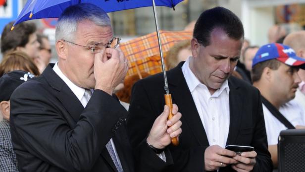 Zwei Männer in Anzügen stehen unter einem blauen Regenschirm, einer schaut auf sein Handy.