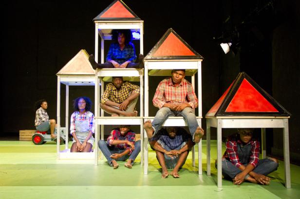 Mehrere Personen sitzen in und unter stilisierten Hausrahmen auf einer Bühne.