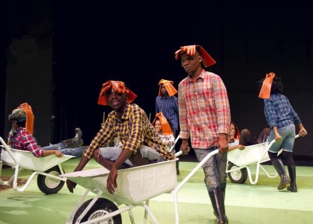 Mehrere Personen mit orangefarbenen Handschuhen auf dem Kopf sitzen oder stehen in Schubkarren auf einer Bühne.