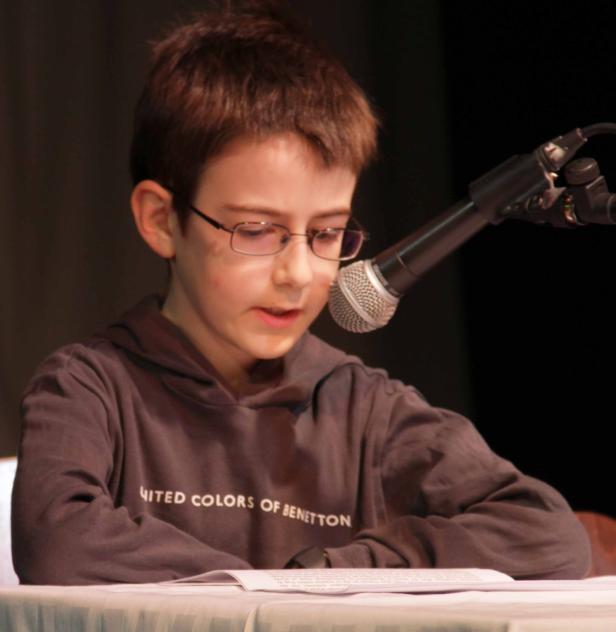 Ein Junge mit Brille liest an einem Mikrofon von einem Blatt Papier ab.