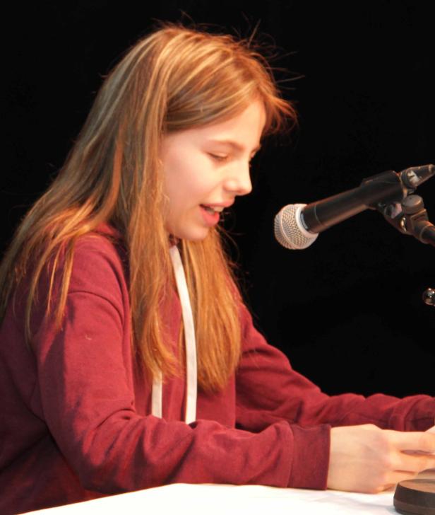 Ein junges Mädchen liest an einem Mikrofon vor Publikum vor.