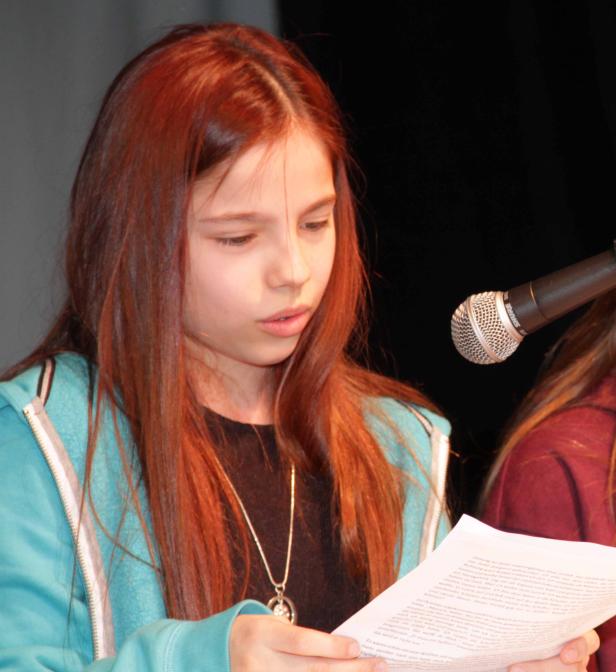 Ein junges Mädchen mit roten Haaren liest einen Text vor einem Mikrofon.