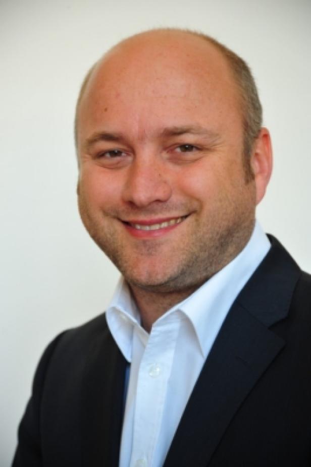 Ein lächelnder Mann in Anzug und Hemd vor einem hellen Hintergrund.