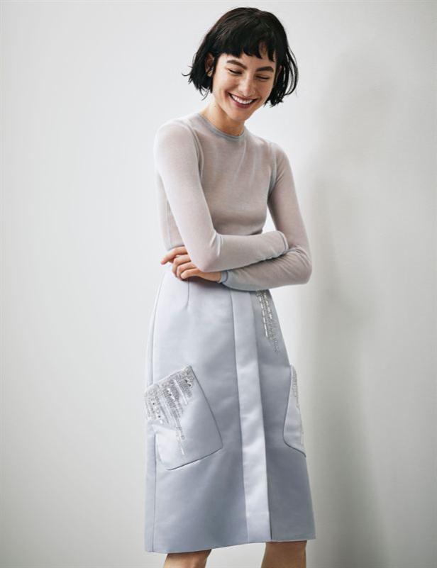 Eine lächelnde Frau in einem hellblauen Rock mit verzierten Taschen und einem langärmligen Oberteil.