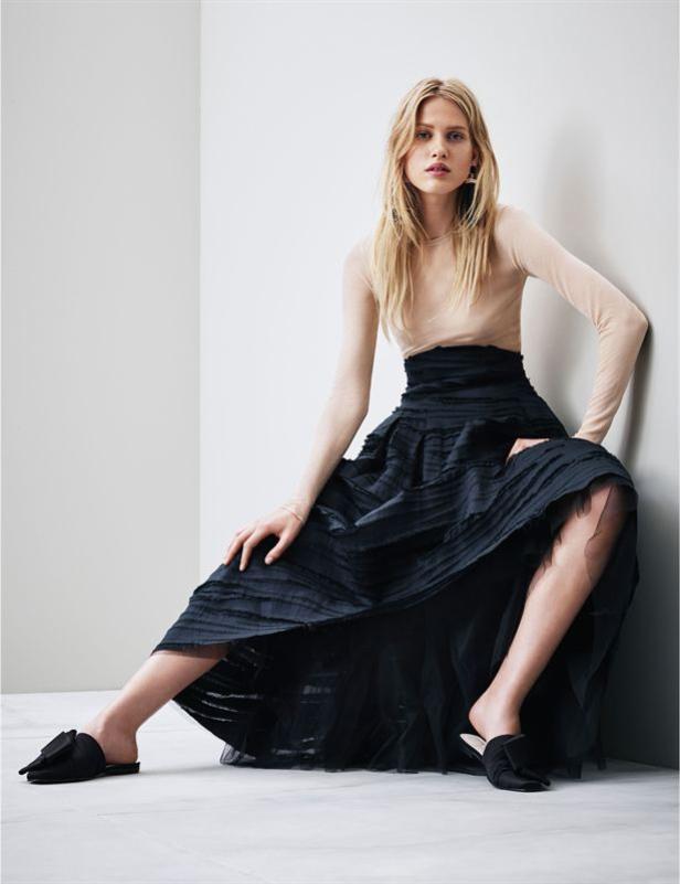 Eine blonde Frau posiert in einem langen, schwarzen Rock und einem hellen Oberteil vor einer Wand.