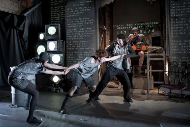 Auf einer Bühne ziehen drei Schauspieler aneinander, während ein Mann mit Ghettoblaster auf einer Leiter sitzt. Im Hintergrund sind beschriftete Wände und Fernseher zu sehen.