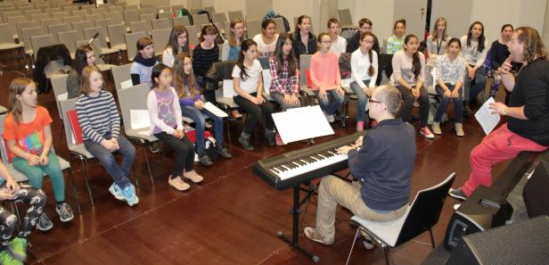 Ein Jugendchor probt mit einem Pianisten und einem Dirigenten in einem Konzertsaal.
