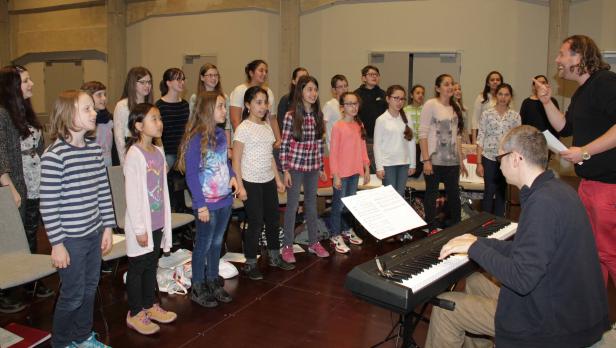 Ein Kinderchor singt unter der Leitung eines Dirigenten, begleitet von einem Pianisten.