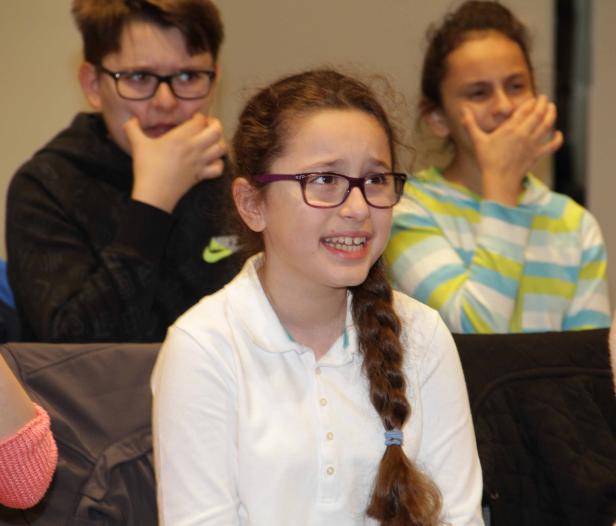 Ein junges Mädchen mit Zopf und Brille lächelt, während andere Personen im Hintergrund sitzen.