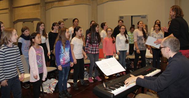 Ein Kinderchor probt mit einem Pianisten und einem Chorleiter.