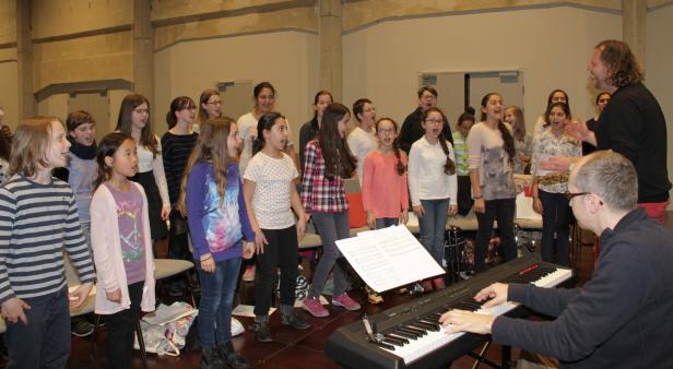 Ein Kinderchor singt unter der Leitung eines Dirigenten, der Klavier spielt.