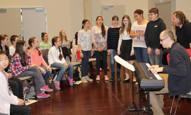 Ein Jugendchor probt mit einem Mann am Keyboard in einem Raum.