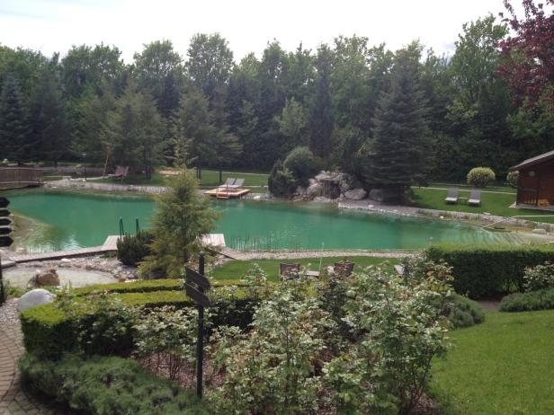 Ein grüner See mit Liegestühlen und einem kleinen Wasserfall in einer parkähnlichen Anlage.