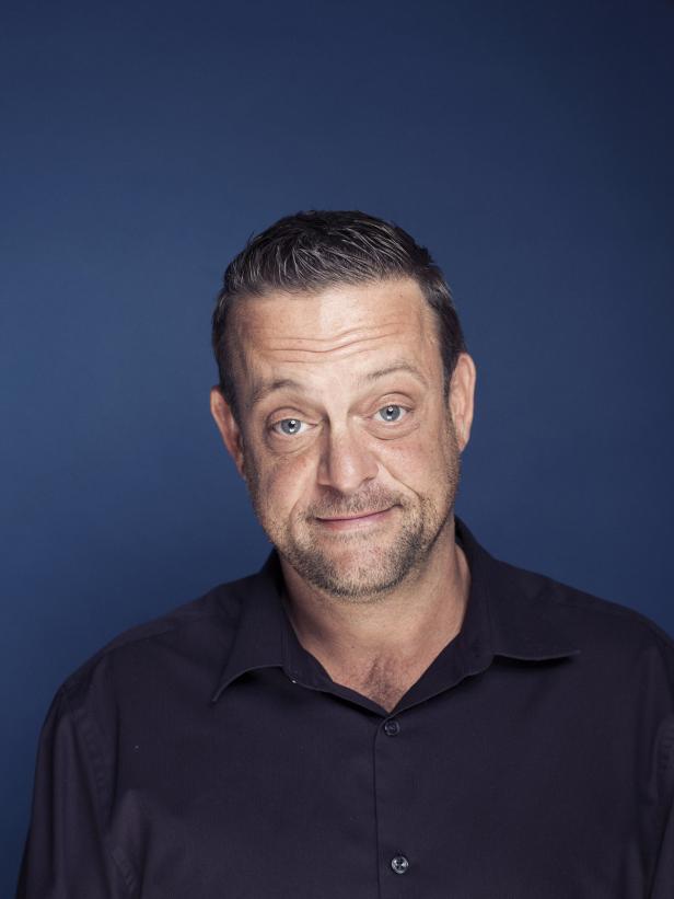 Ein Mann mit Bart und blauem Hemd lächelt vor einem blauen Hintergrund.