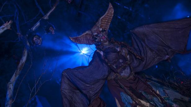 Eine Person in einem Fledermauskostüm steht vor einem blauen Hintergrund mit Fledermäusen an Ästen.