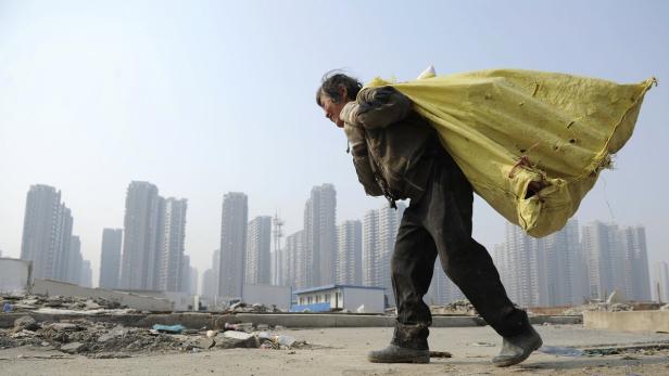 Eine ältere Person trägt einen schweren Sack vor einer Skyline aus Hochhäusern.