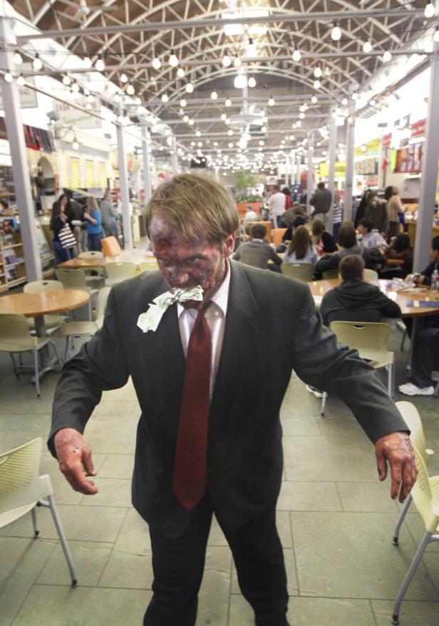 Ein als Zombie verkleideter Mann im Anzug steht in einer Cafeteria und hat Geldscheine im Mund.