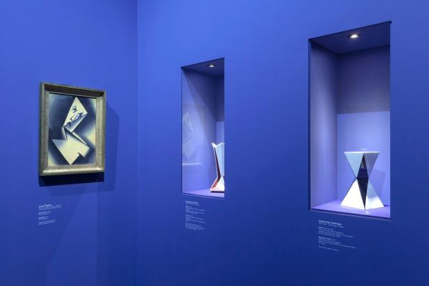 Eine Kunstausstellung mit einem Gemälde und zwei Vasen in Vitrinen vor blauem Hintergrund.