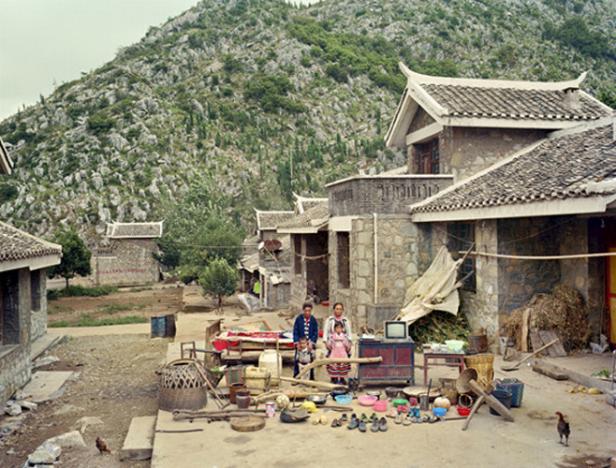 Eine Familie steht vor ihrem Haus in einem chinesischen Dorf, im Hintergrund ein bewaldeter Berg.