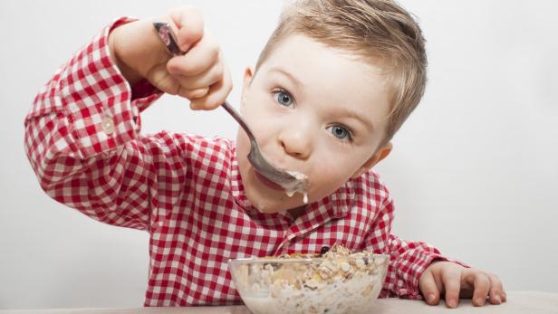 Ein Junge im rot-weiß karierten Hemd isst Müsli mit einem Löffel.