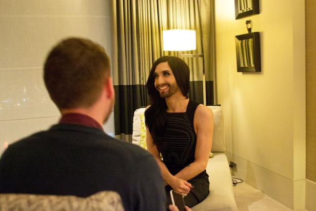 Conchita Wurst sitzt in einem Raum und lächelt einen Mann an.