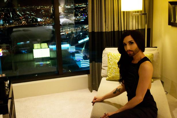 Conchita Wurst sitzt in einem eleganten Zimmer mit Blick auf eine beleuchtete Stadt.