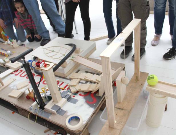 Eine selbstgebaute Murmelbahn aus Holz mit verschiedenen Hindernissen und einem gelben Tennisball am Ende.