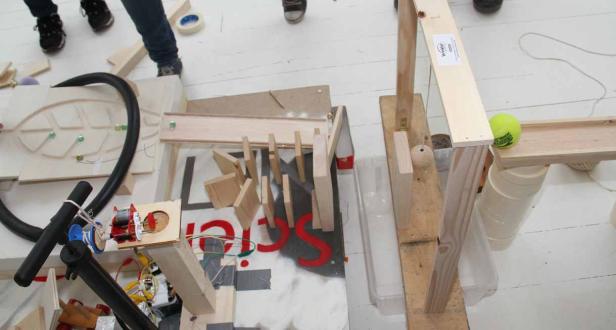 Eine selbstgebaute Murmelbahn mit verschiedenen Hindernissen und einer gelben Tennisball am Ende.