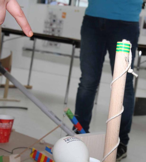 Ein Finger zeigt auf eine selbstgebaute Vorrichtung mit einem Holzzylinder und einer Kugel.