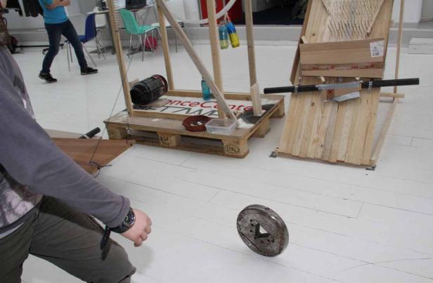 Ein Mann beobachtet, wie ein Metallteil in einem Raum mit verschiedenen Holzkonstruktionen zu Boden fällt.