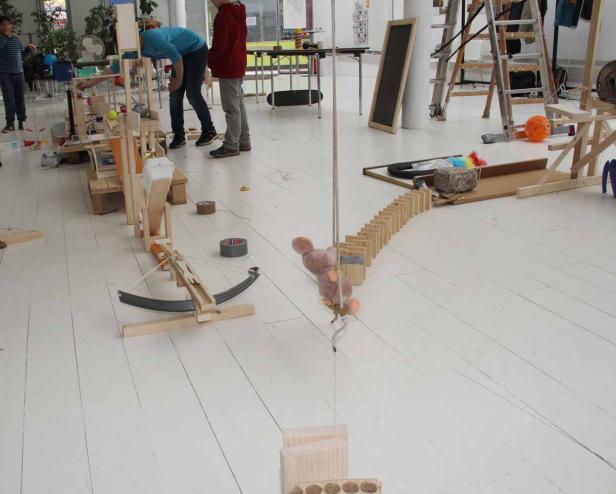 Eine komplexe Kettenreaktion aus Holzbauteilen und einem Teddybär in einem hellen Raum.