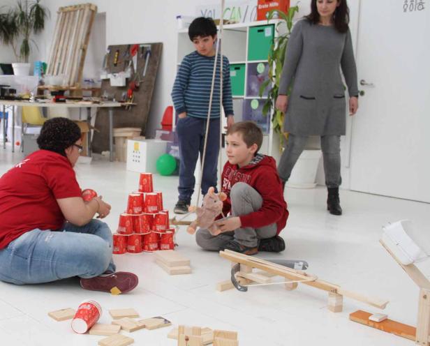Kinder spielen mit Bechern, Holzklötzen und einer selbstgebauten Balliste in einem hellen Raum.