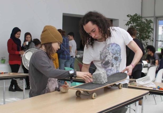 Zwei Personen hantieren mit einem Skateboard und einem Stein auf einem Tisch.