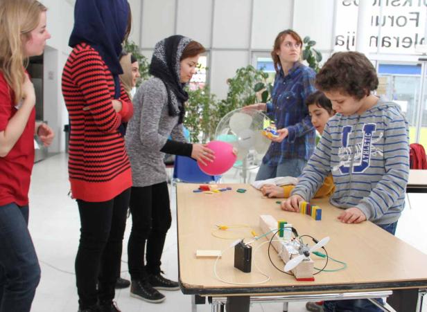 Eine Gruppe von Menschen, darunter Kinder, experimentieren mit einem Ventilator und einem Ballon an einem Tisch.