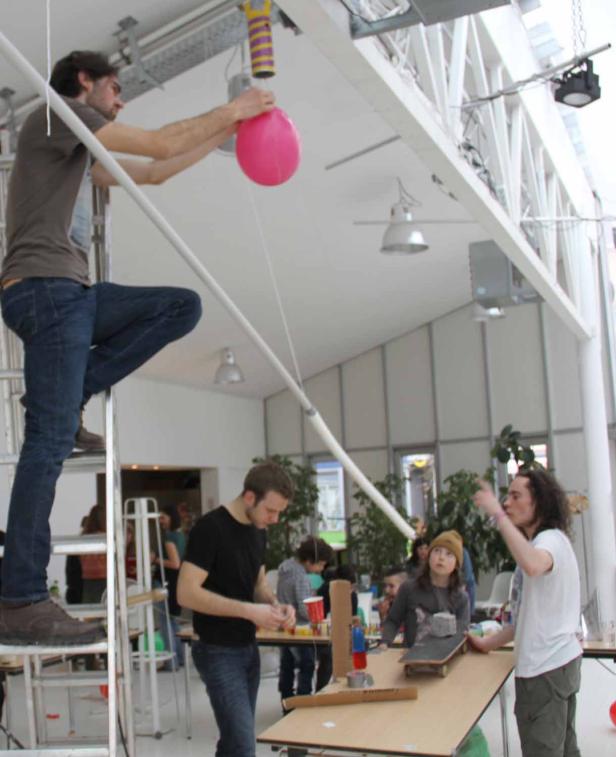 In einem hellen Raum bereiten mehrere Personen ein Experiment mit einem Ballon und einer Rampe vor.