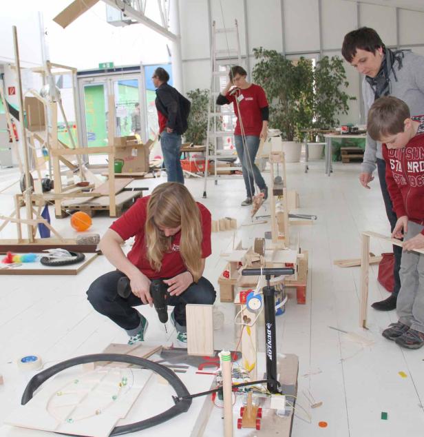 Eine Frau bohrt in einem hellen Raum an einer Murmelbahn, während andere Personen zusehen.