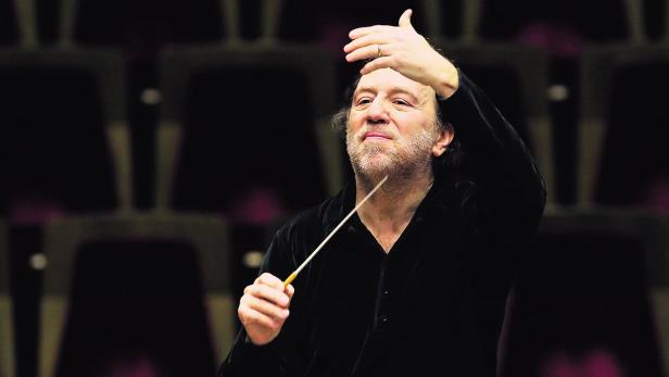 Ein Dirigent mit erhobener Hand und Taktstock vor einem dunklen Hintergrund.