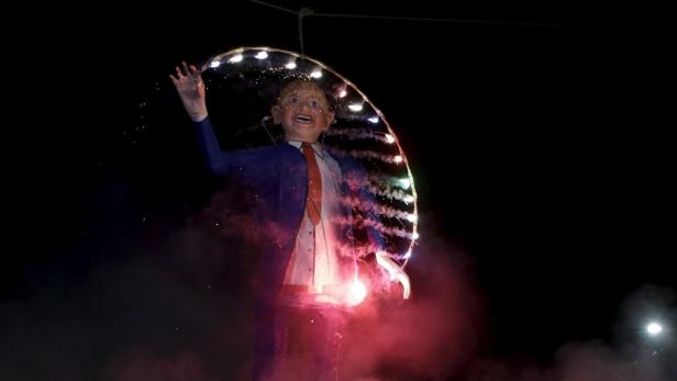Eine Donald Trump-Figur steht inmitten von Feuerwerk und Rauch.
