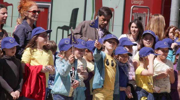 Eine Gruppe Kinder mit EU-Fahnen und blauen Kappen mit EU-Sternen bei einer Veranstaltung.