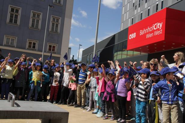 Eine Gruppe Kinder mit EU-Fahnen und blauen Kappen vor der ÖBB BahnhofCity Wien West.