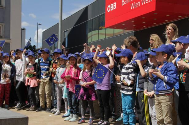 Eine Gruppe Kinder mit EU-Fahnen und -Hüten vor dem ÖBB Bahnhof Wien West.