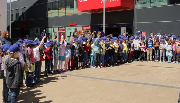 Eine Gruppe Kinder mit EU-Fahnen und blauen Kappen vor dem Bahnhof Wien West.