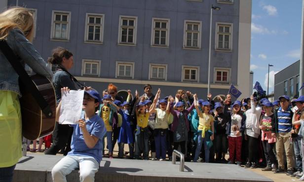 Eine Gruppe Kinder mit EU-Flaggen singt mit musikalischer Begleitung vor einem Gebäude.