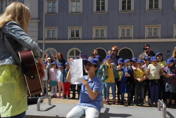 Eine Gruppe Kinder mit EU-Fahnen singt vor einem Gebäude, begleitet von einer Gitarristin.