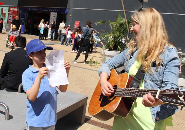 Eine Frau spielt Gitarre, während ein Junge mit Europahut einen Text liest.