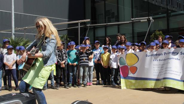 Eine Frau spielt Gitarre vor einer Gruppe von Kindern, die ein Schild der European Primary School halten.