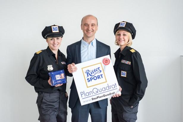 Zwei Frauen in Uniform und ein Mann präsentieren eine Tafel von Ritter Sport und ein Schild von „PlanQuadrat“.