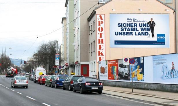 Eine Straße in einer Stadt mit parkenden Autos und einem großen Werbeplakat für die Hypo Oberösterreich.