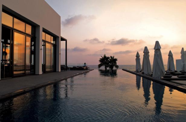 Ein Infinity-Pool spiegelt den Himmel bei Sonnenuntergang wider, mit Palmen und Sonnenschirmen am Meer.