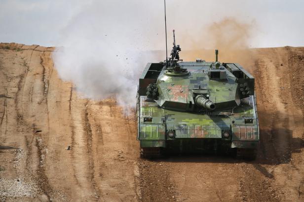 Ein getarnter Panzer fährt einen Hügel hinauf, Staub aufwirbelnd.